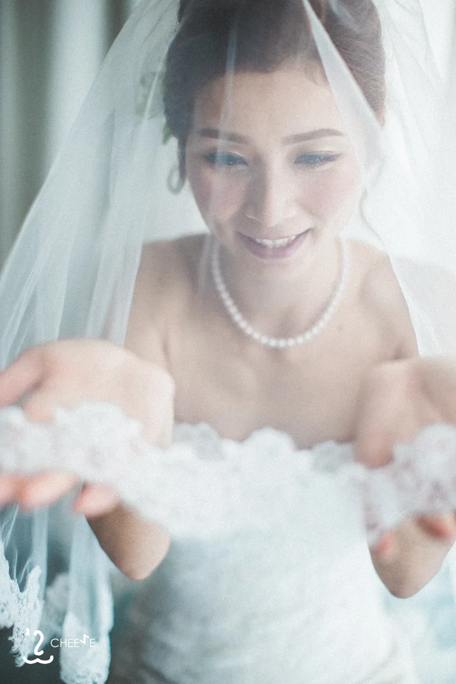 頭紗下的新娘低頭微笑,雙手提起蕾絲婚紗細節